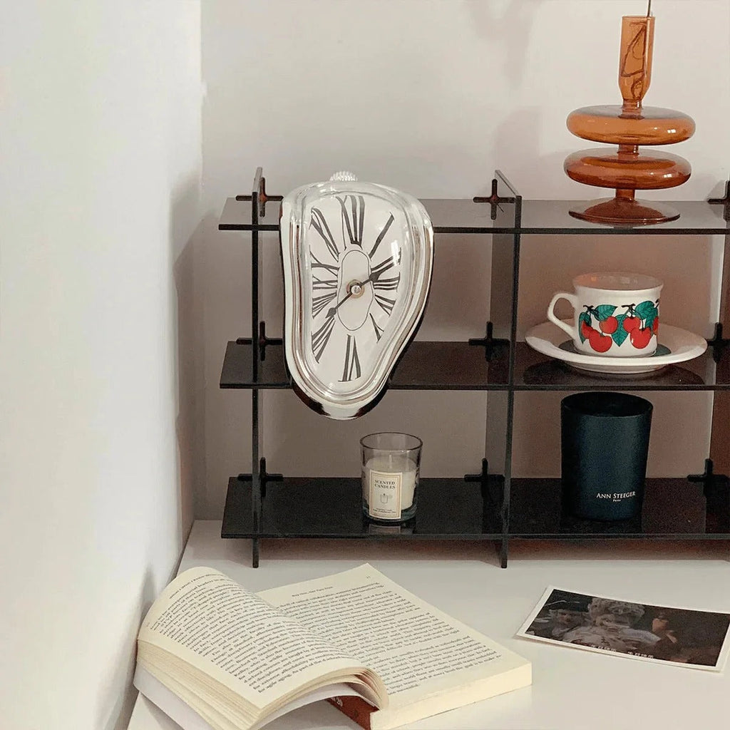 Silver melting clock draped over the front edge of a black steel shelving unit, beside a lit candle, a cherry-print cup, an open book and a postcard