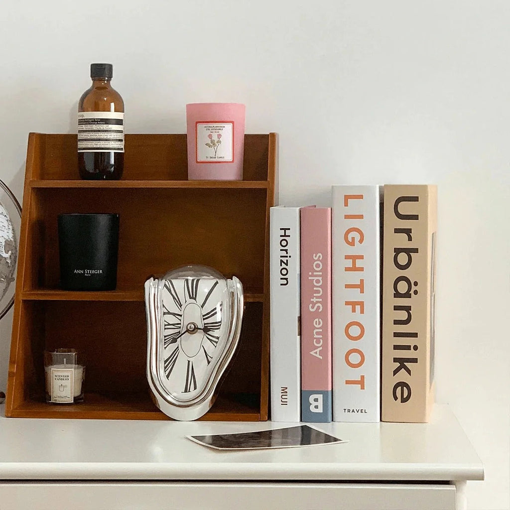 Silver melting clock sitting inside a wooden cubby shelf, beside a brown bottle, a pink candle and a row of art books