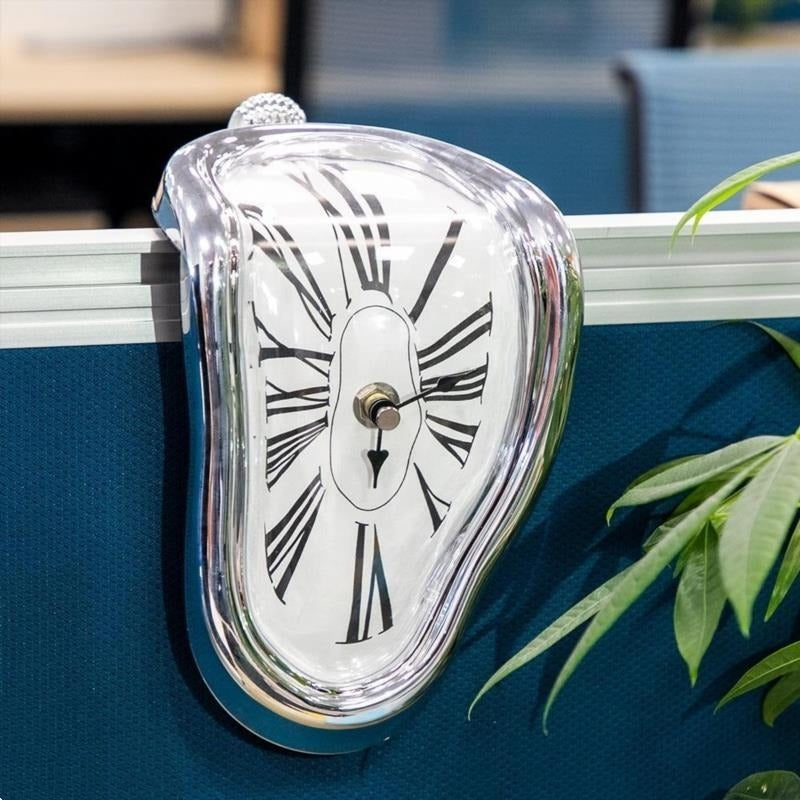 Silver melting clock hanging over the edge of a teal desk divider in a real office, with a plant frond at the right of the frame
